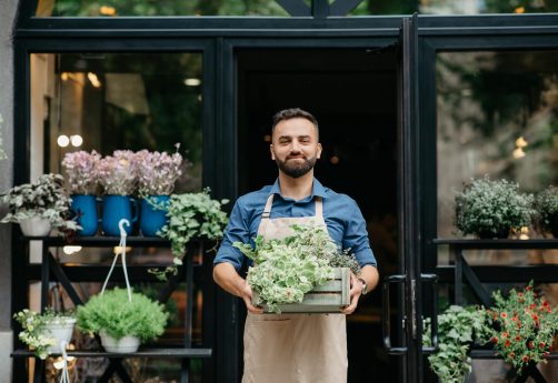 Small business owner works in flower shop and customer service. Confident smiling young bearded european male in apron carries wooden box with plants, on shop or eco cafe background, empty space
